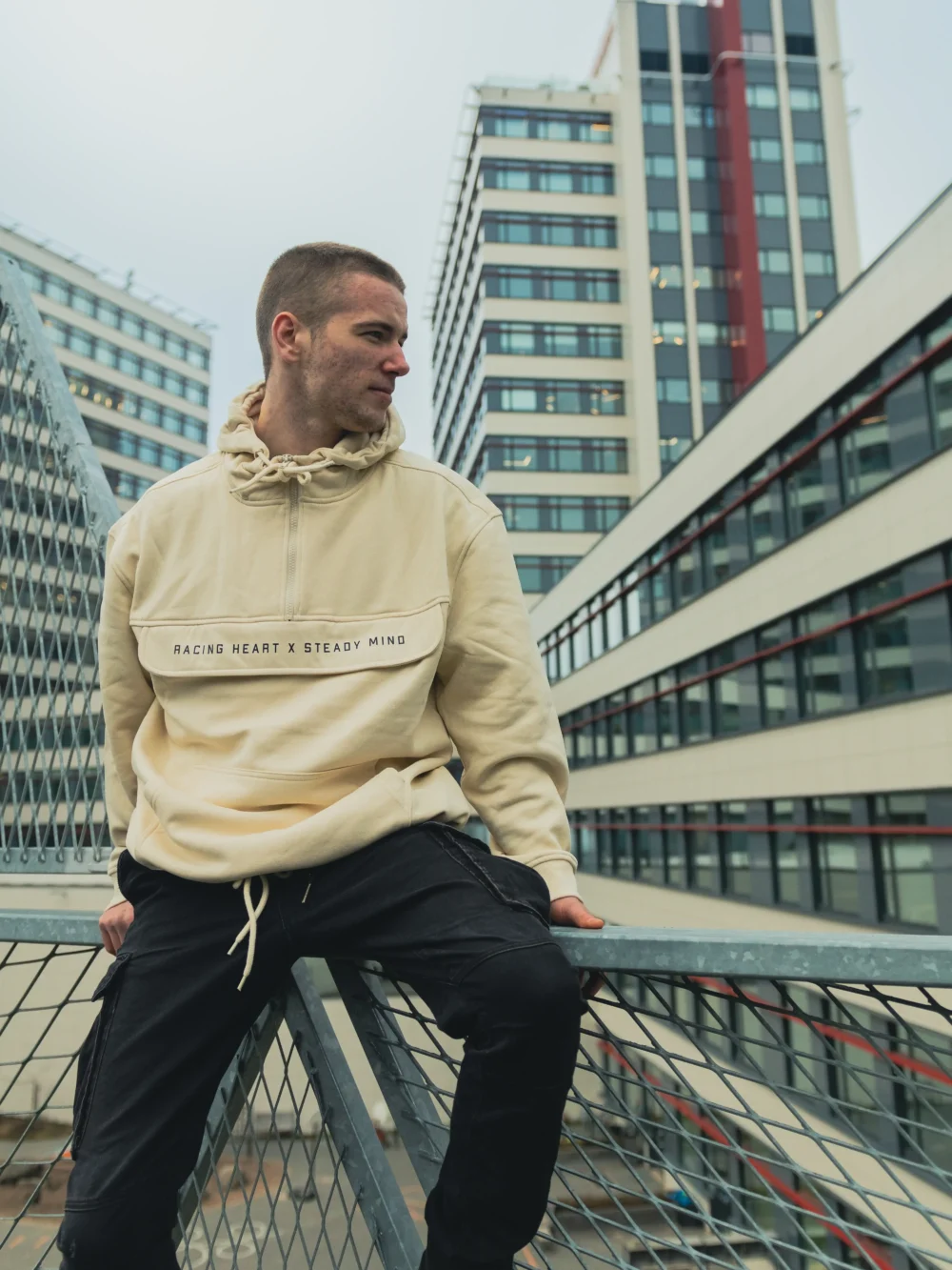 A model sitting in an urban environment wearing the beige RHSM MVMT Hoodie, showcasing its relaxed fit for everyday streetwear.