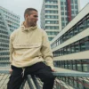 A model sitting in an urban environment wearing the beige RHSM MVMT Hoodie, showcasing its relaxed fit for everyday streetwear.