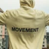 Model wearing the beige RHSM MVMT Hoodie in an urban setting, with a clear view of the "MOVEMENT" back design.