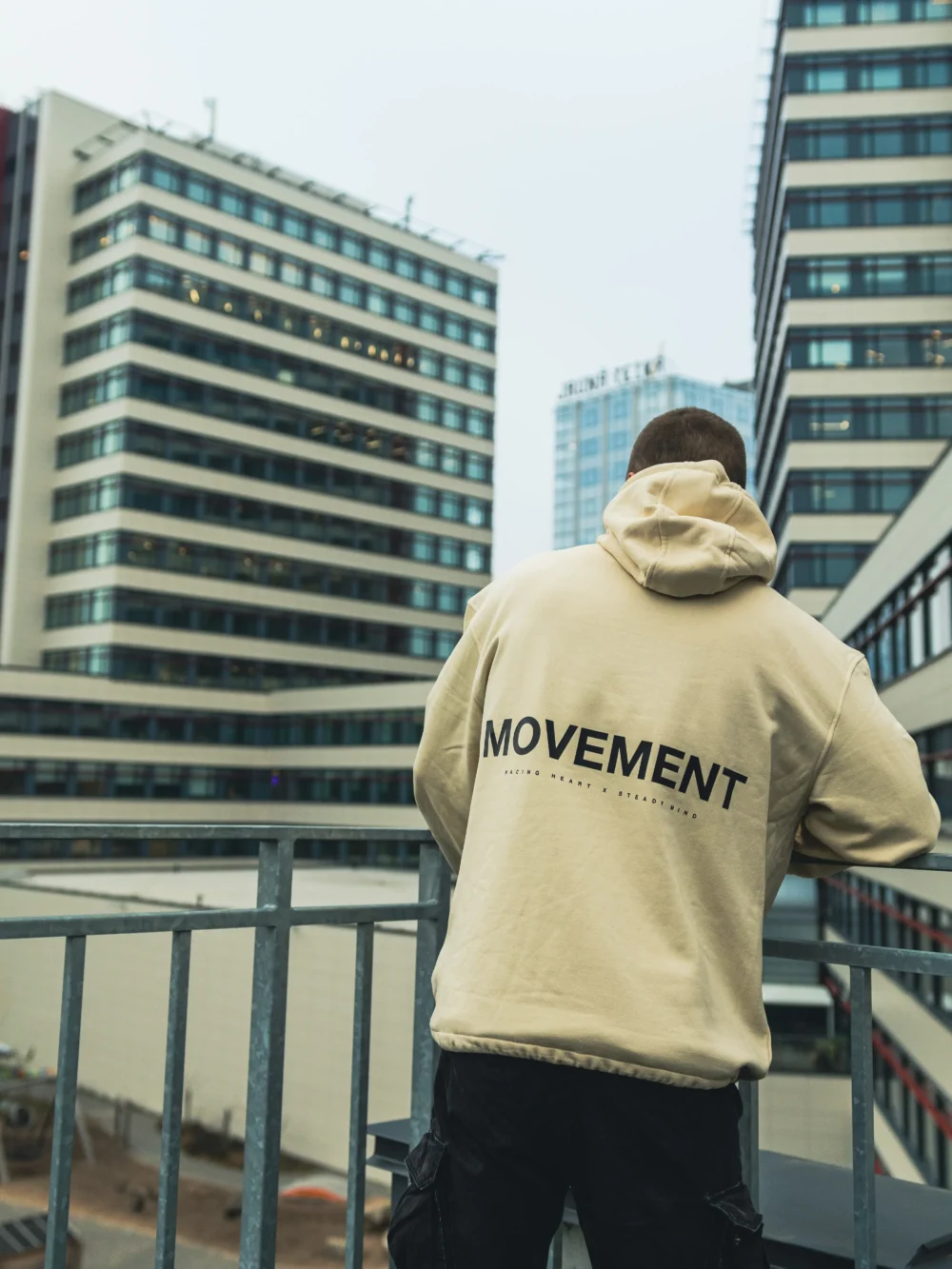Back view of the beige RHSM MVMT Hoodie with its "MOVEMENT" graphic, set against a background of high-rise city buildings.
