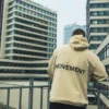Back view of the beige RHSM MVMT Hoodie with its "MOVEMENT" graphic, set against a background of high-rise city buildings.