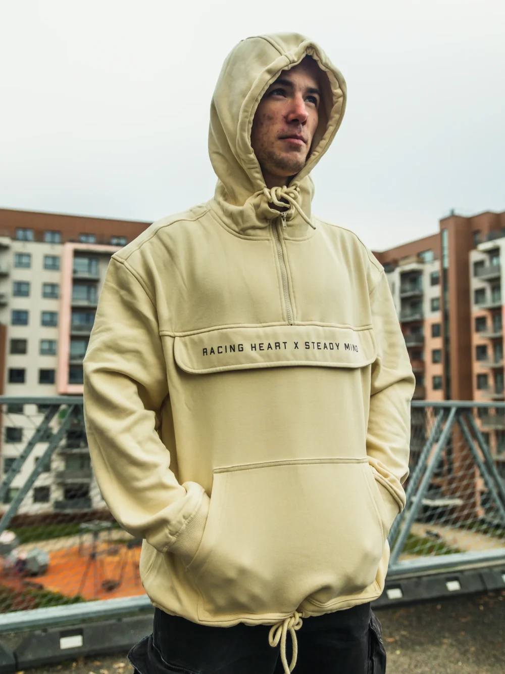 Model in an urban setting wearing the beige RHSM MVMT Hoodie with the hood on, secured by its thick drawstrings.