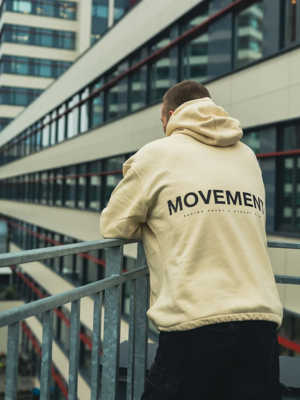 A detailed close-up of the "MOVEMENT" back graphic on the beige RHSM MVMT Hoodie, with an urban building background.