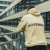 A detailed close-up of the "MOVEMENT" back graphic on the beige RHSM MVMT Hoodie, with an urban building background.