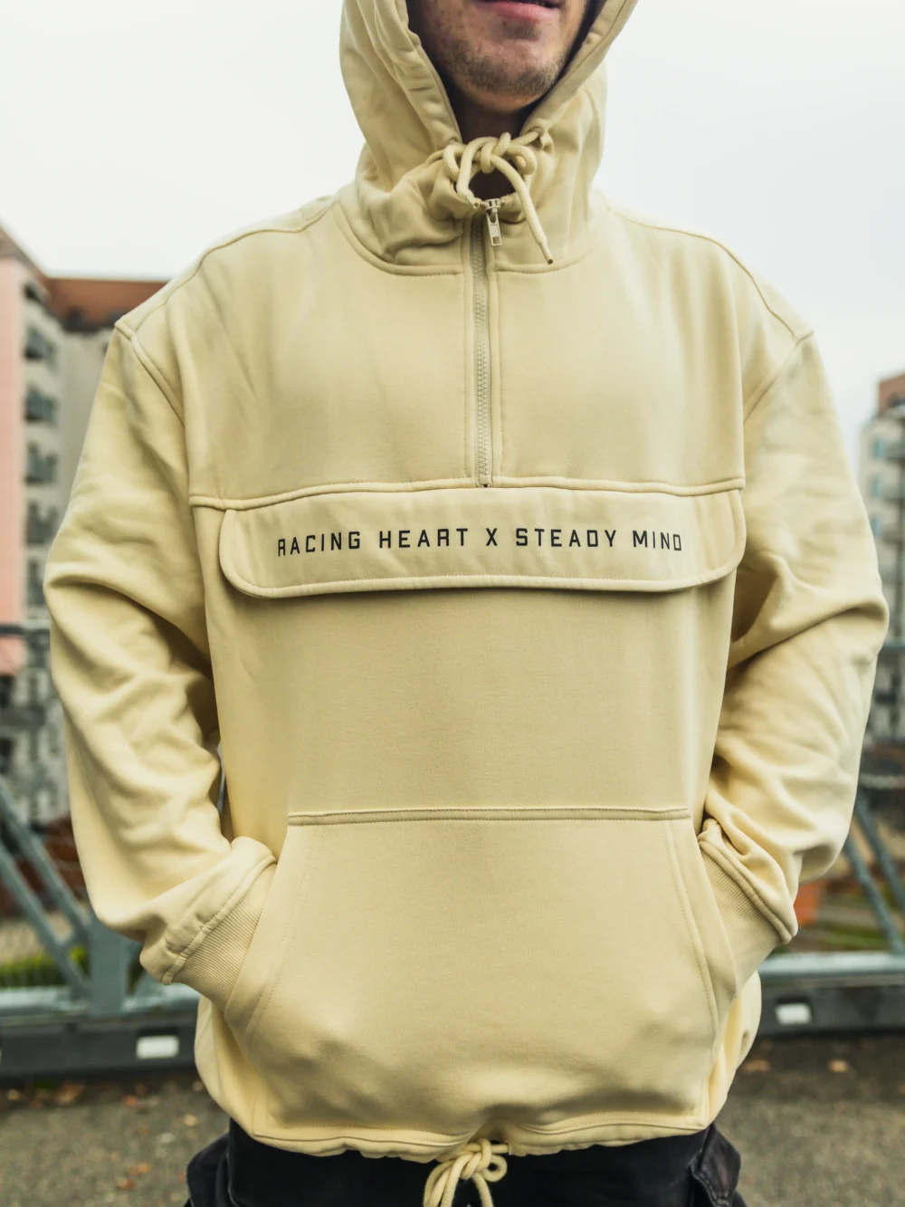 A close-up of a model with hands in the spacious front pouch of the beige RHSM MVMT Hoodie.