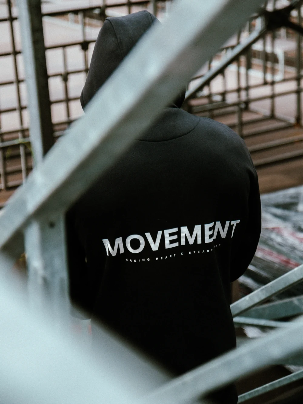 Back view of a model wearing the black RHSM MVMT Hoodie in stadium stands, showcasing the minimalist "MOVEMENT" graphic.