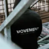 Back view of a model wearing the black RHSM MVMT Hoodie in stadium stands, showcasing the minimalist "MOVEMENT" graphic.