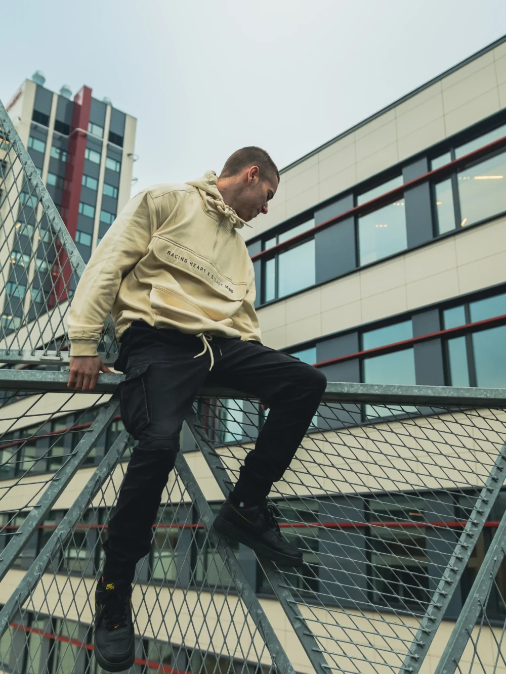 A stylish shot of a model sitting and wearing the beige RHSM MVMT Hoodie in a well-composed urban architectural setting.