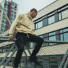 A stylish shot of a model sitting and wearing the beige RHSM MVMT Hoodie in a well-composed urban architectural setting.