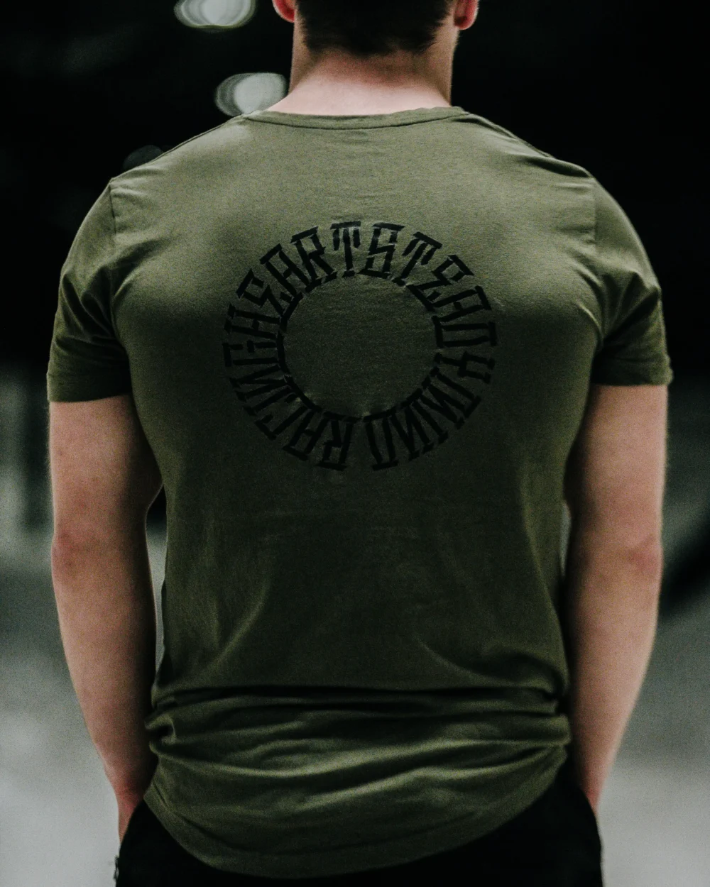 Back view of a model wearing the khaki RHSM Long Fit Tee V2 outdoors, with a clear view of the back graphic.