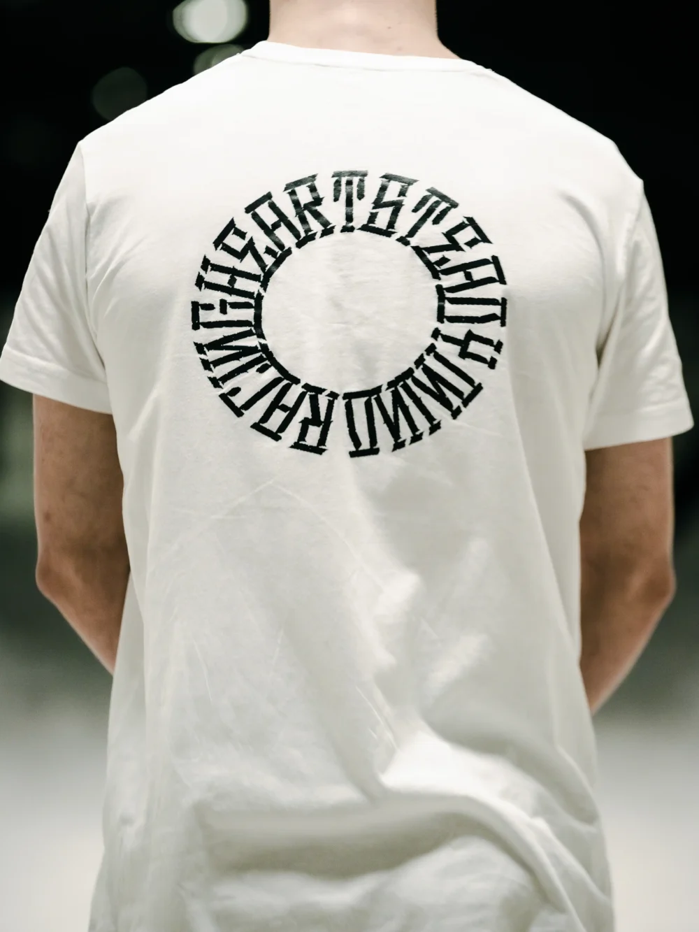 Back view of a model wearing the white RHSM Long Fit Tee V2 outdoors, with a clear view of the back graphic.