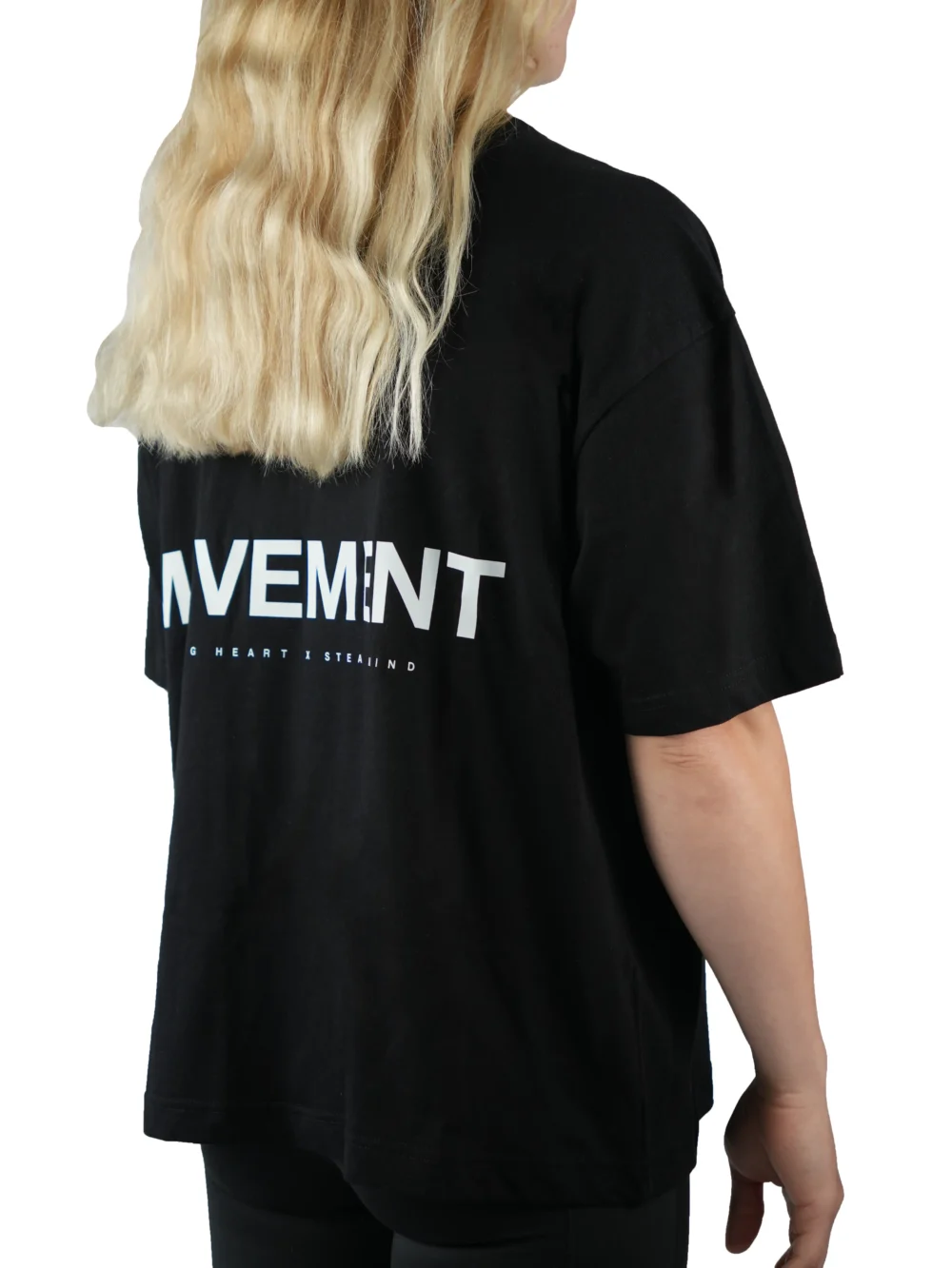 Back view of a female model wearing the black oversized boxy tee, showcasing the white "Movement" graphic and the tee's wide, loose fit.