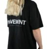 Back view of a female model wearing the black oversized boxy tee, showcasing the white "Movement" graphic and the tee's wide, loose fit.