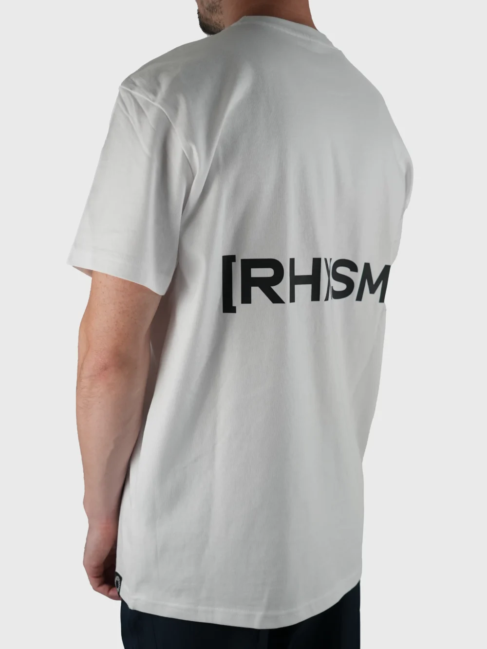 Model wearing the white "[RHXSM]" tee (back view), showcasing the impactful back design and dropped shoulders from the calisthenics clothing brand RHSM.