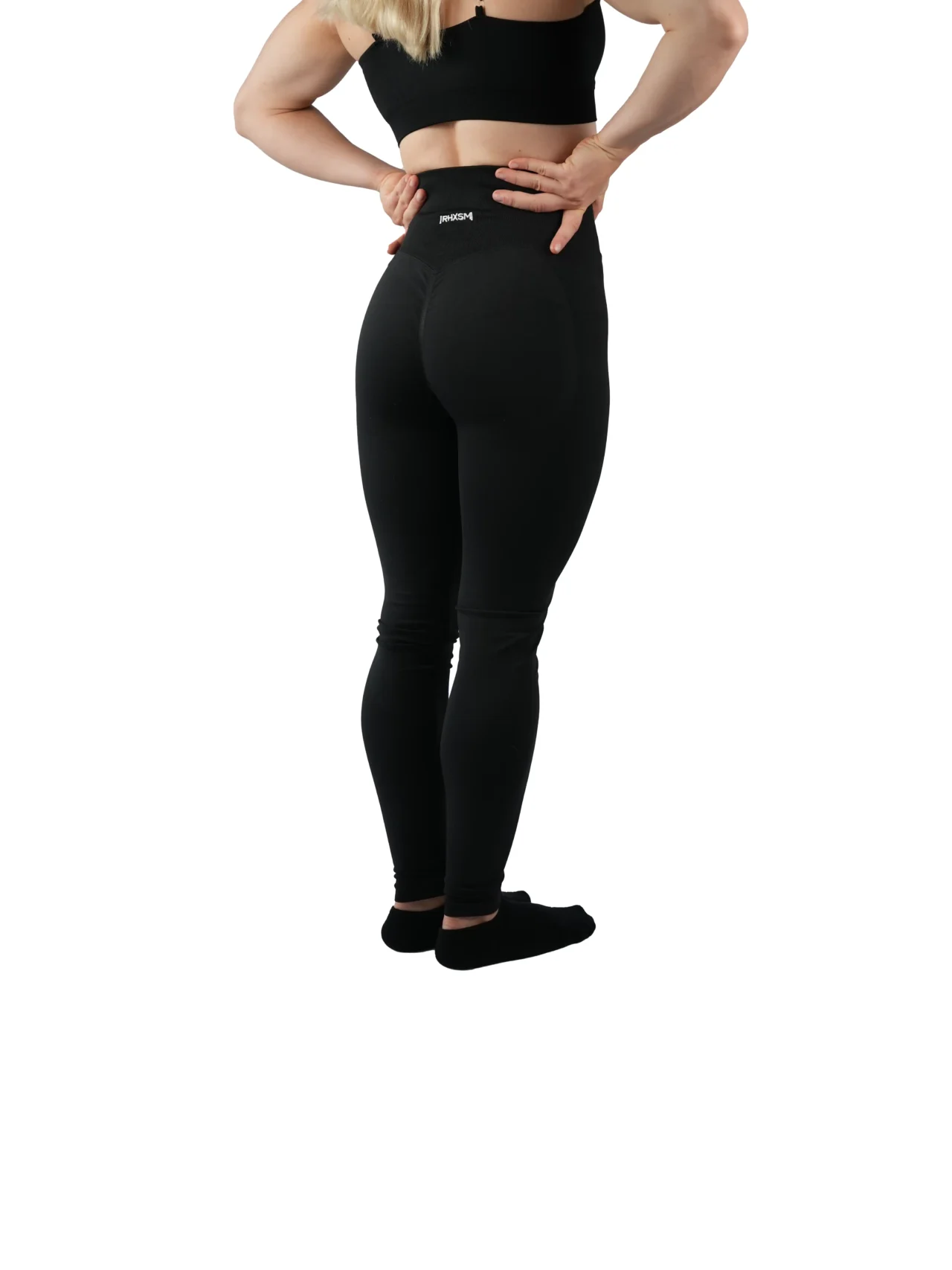 Model wearing the black "[RHXSM]" leggings (back view), showcasing the flattering scrunch detail and subtle embroidered logo from the calisthenics clothing brand RHSM.