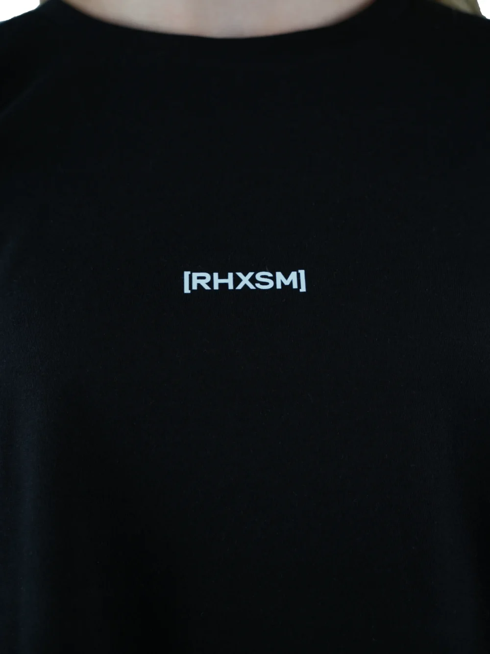 A close-up of the black "[RHXSM]" long sleeve, detailing the subtle front graphic and sewn-in hem label on this piece of performance apparel.