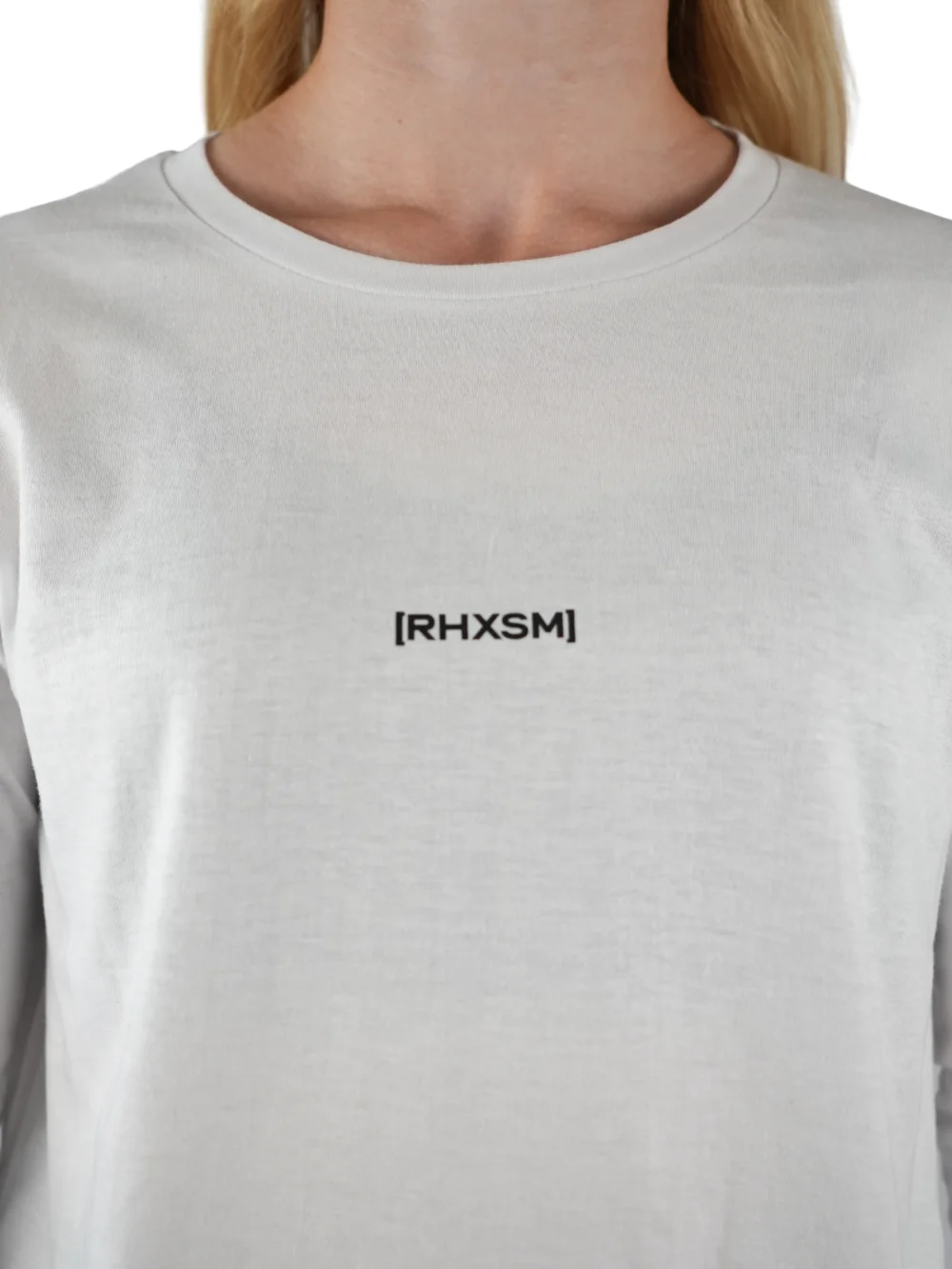 A close-up of the white "[RHXSM]" long sleeve tee, showing the clean front graphic and premium sewn-in label from the calisthenics clothing brand RHSM.