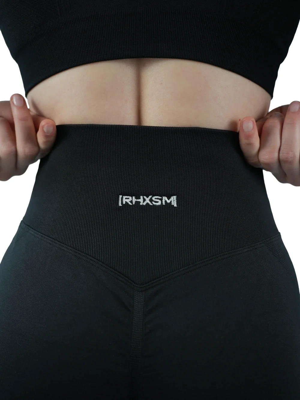 A close-up of the black "[RHXSM]" seamless leggings, detailing the premium embroidered back graphic with its anti-scratch interior backing for comfort.